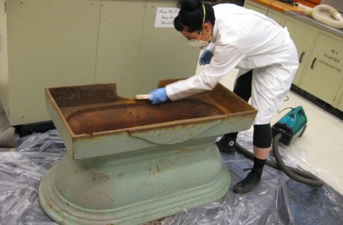 An individual wearing a white lab coat, face mask, and blue gloves uses a stiff brush to clean an upside-down water trough upturned on clear plastic.