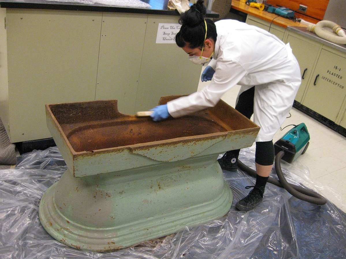 An individual wearing a white lab coat, face mask, and blue gloves uses a stiff brush to clean an upside-down water trough upturned on clear plastic.