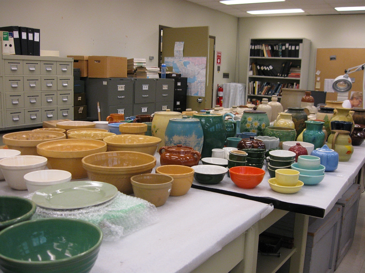 A variety of Medalta pottery items including jars, bowls, and pitchers, arranged across two tables in a museum storage room.