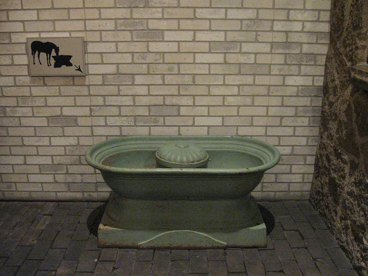 A water trough with a fountain-like piece in the centre, placed against a brick wall near a sign showing a horse drinking from a similar shaped trough, and an arrow pointing towards this one.