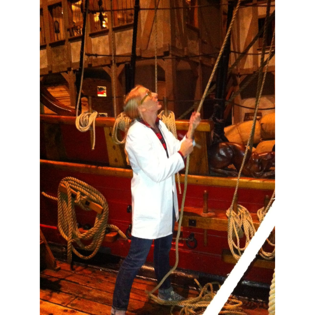 A woman wearing a white lab coat stands on the deck of a wooden sailing vessel, holding a rope taught as she looks up.