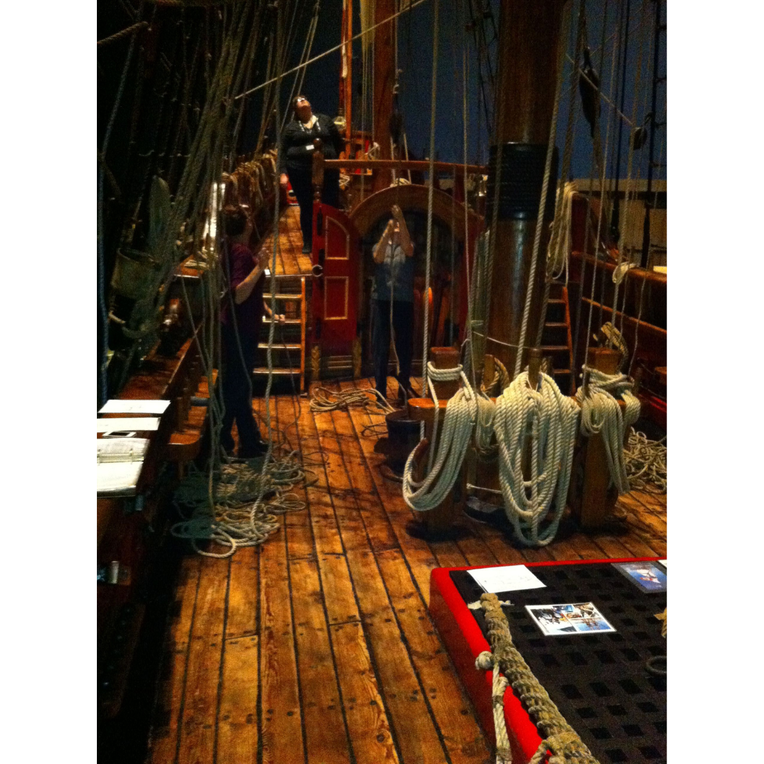 Three individuals standing on the deck of a wooden sailing vessel. All are looking up, as one pulled on a rope threaded through a pulley.