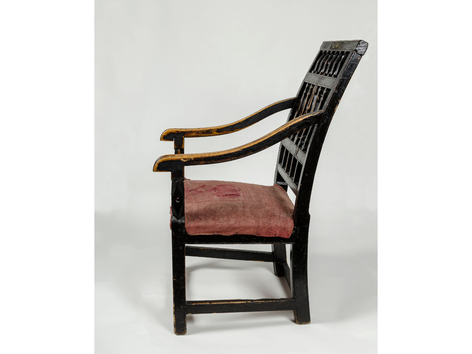 A dark-coloured dining chair chair from the side. The back curves up and backwards, and the two arms slant down towards the front, with visible wear on the top of them from use. The seat is upholstered in a worn red fabric.