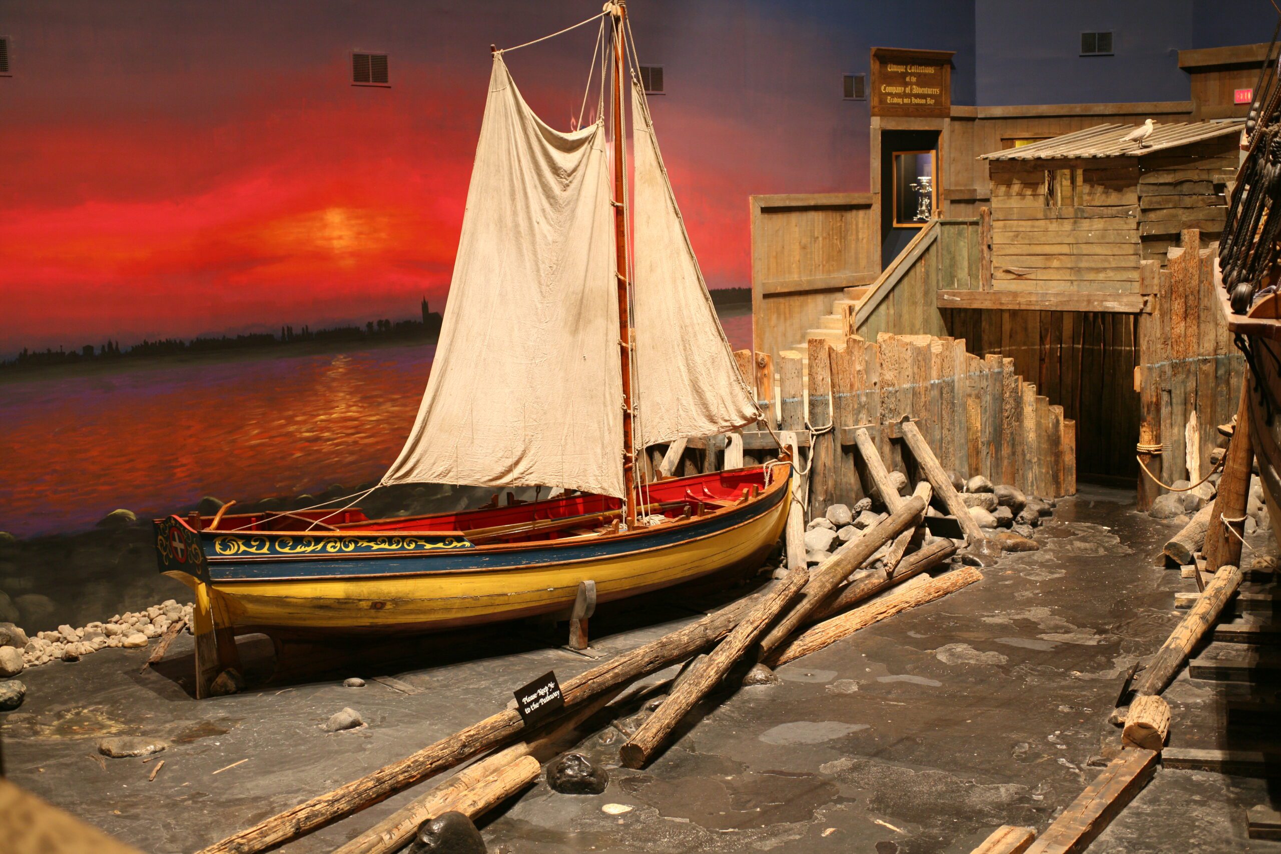 A small wooden sailing vessel, a shallop, on display in a museum gallery, in front of a wall mural depicting a sunset that shallop's white sails are raised.