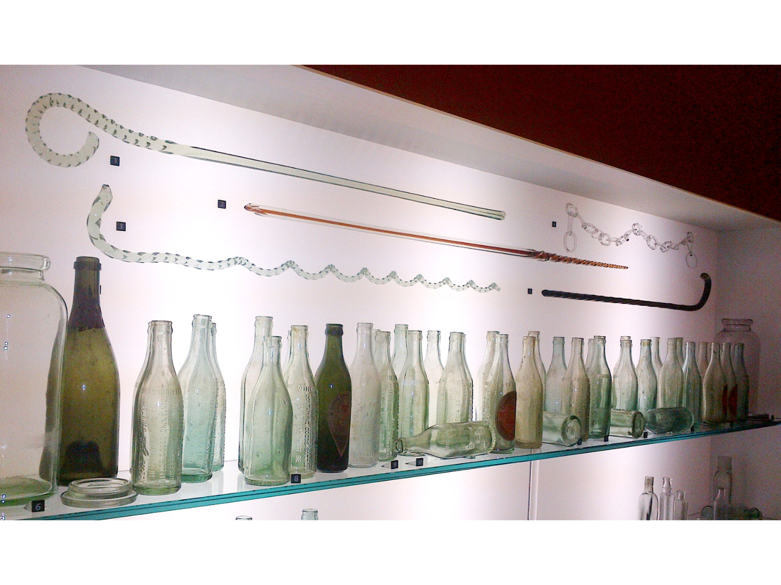 An illuminated display case with a shalf of glass bottles and a number of glass canes suspended above.