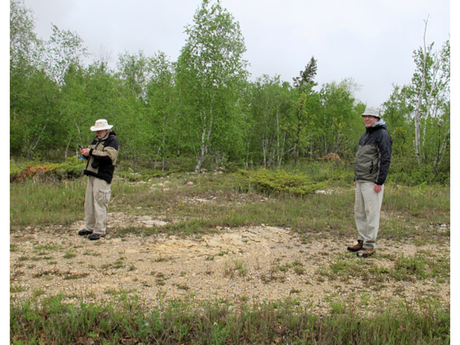 Two individuals standing on a rough gravel path next to a grassy space and some green-leaved trees. Ons of the individuals is holding and examining a handheld device.