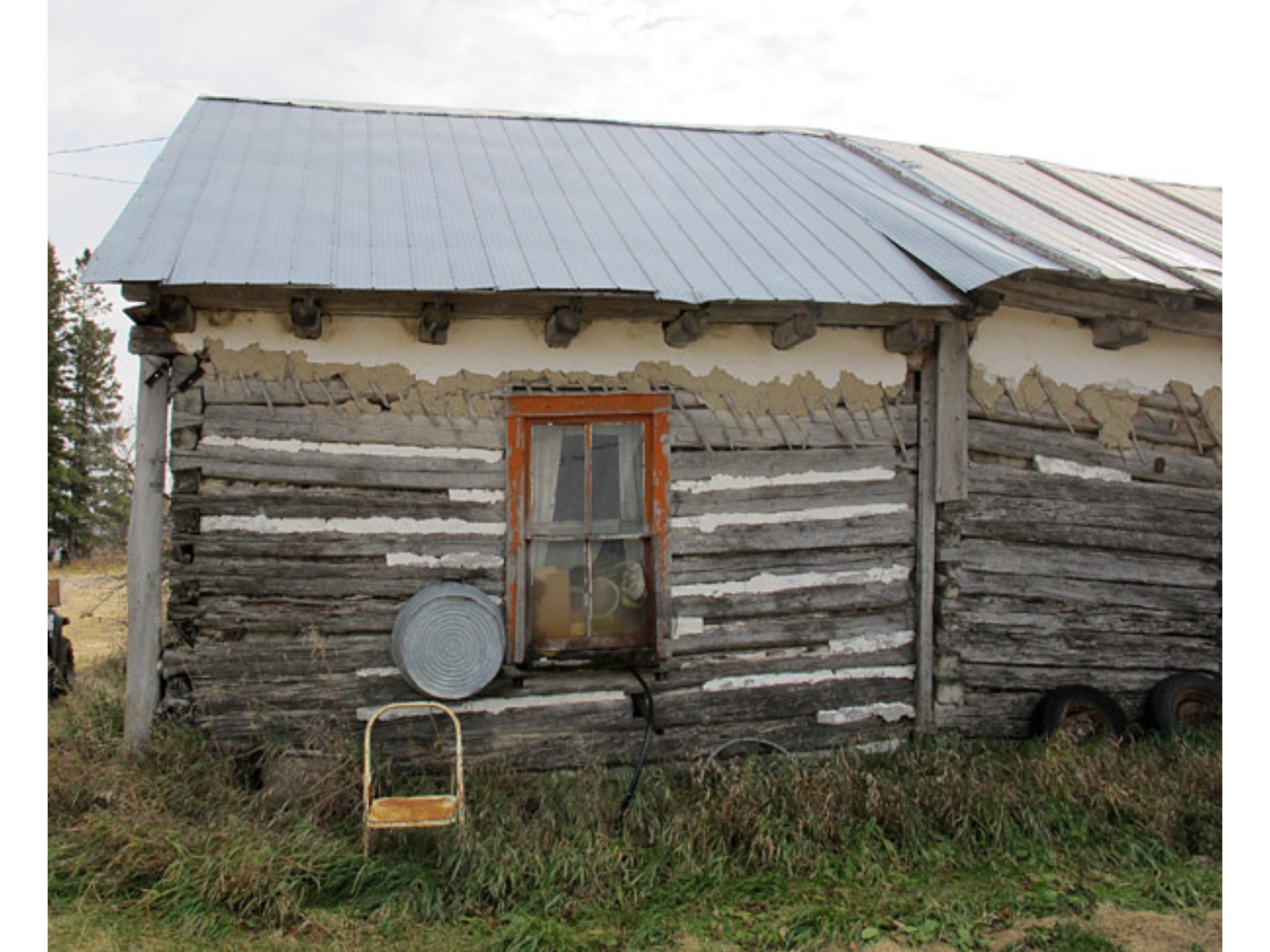 An old farm building with a folding chair and metal washbasin outside next to a window with peeling orange trim and drawn curtains on the other side of the fogged glass.