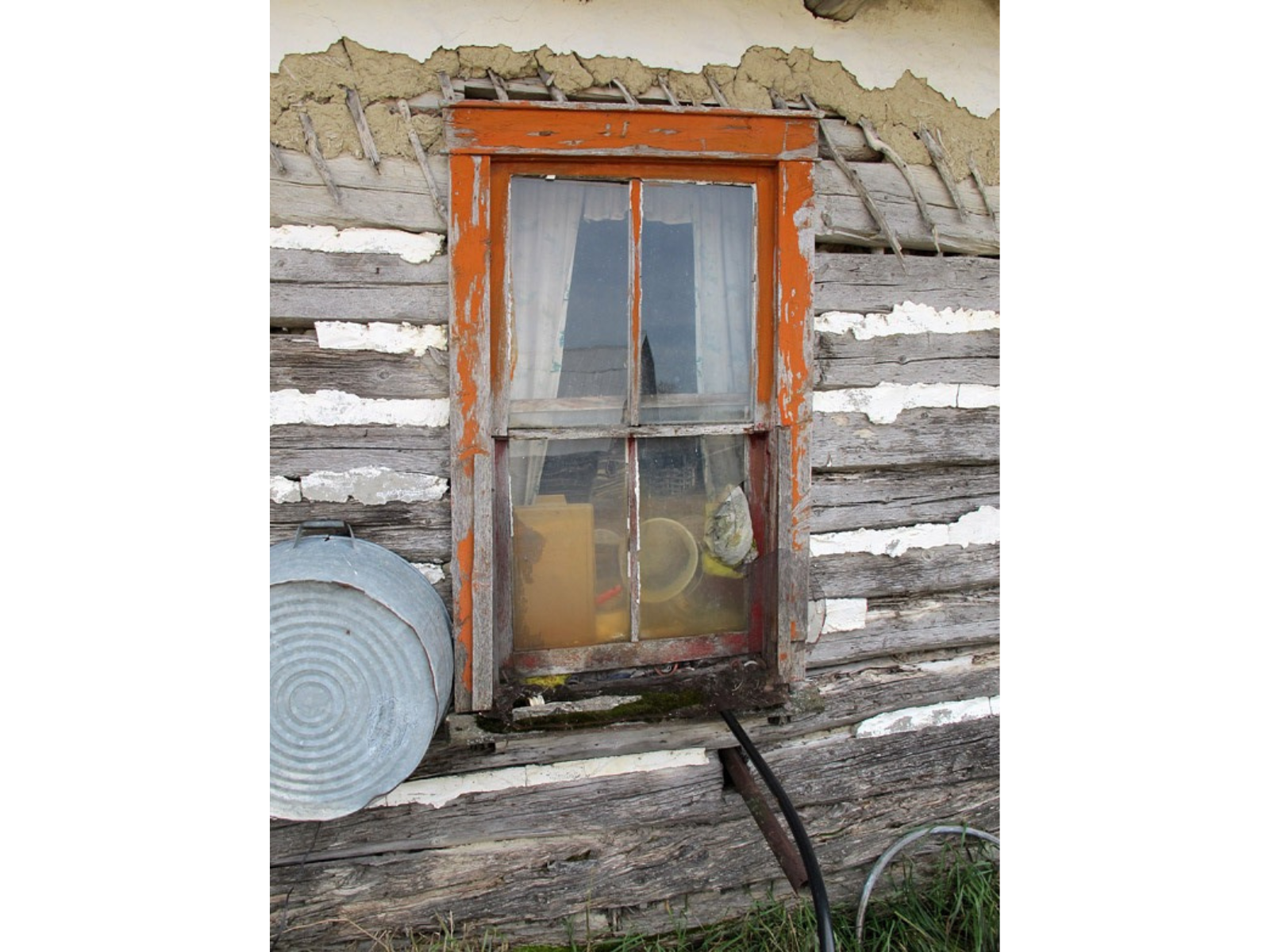 Close-up on the window in the previous image. On the exterior of the old wooden walls hangs a metal washbasin. The window trim is orange and peeling. Inside some clutter is up against he window glass, seen between the drawn curtains.