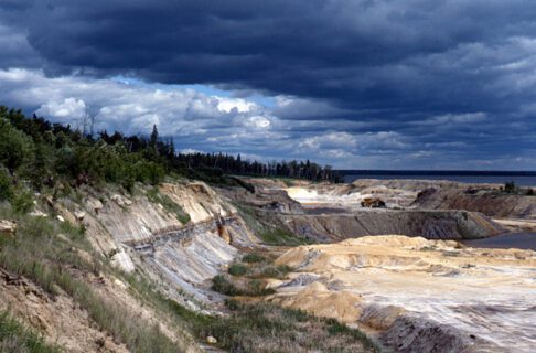 A landscape looking out over the cliffside of a stretch of land leading down to a shoreline. The sky is dramatically lit with thick clouds in shades of light and dark blue.