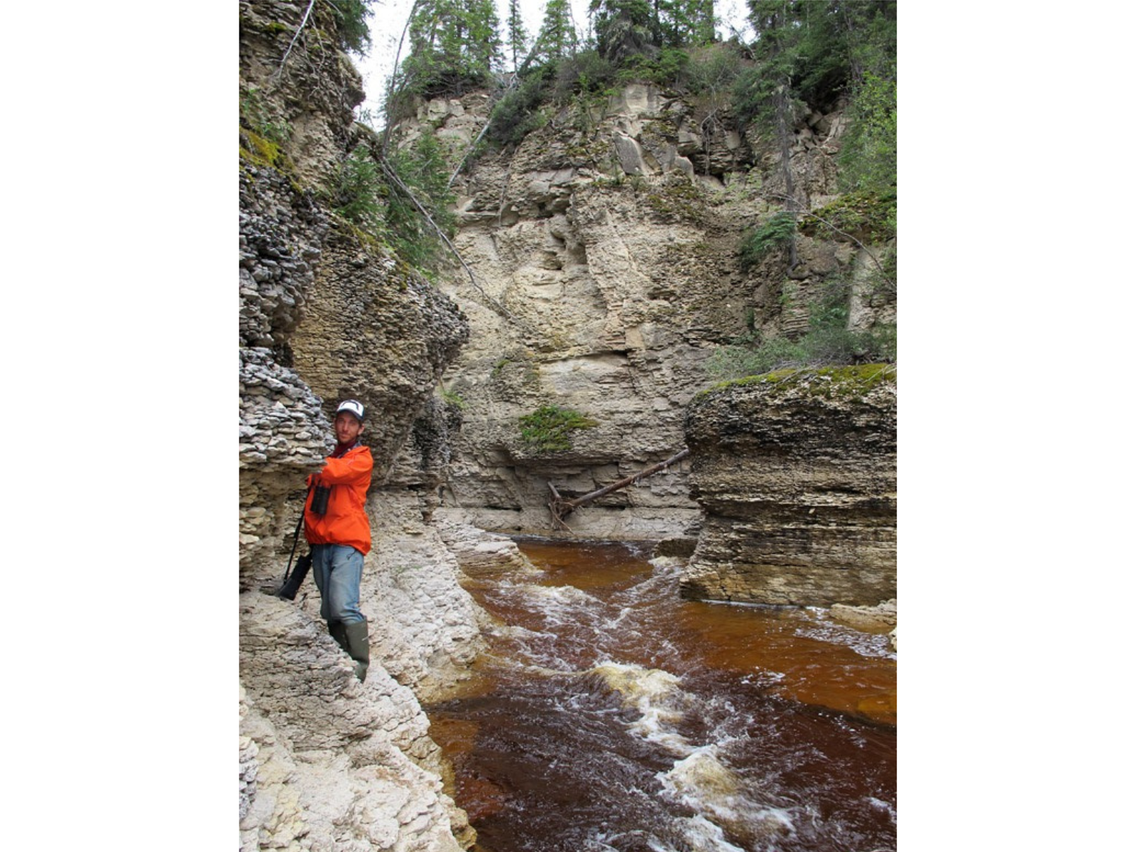 A person wearing an orange jacket standing on a narrow ridge of the cliffside of a steep-sided river.