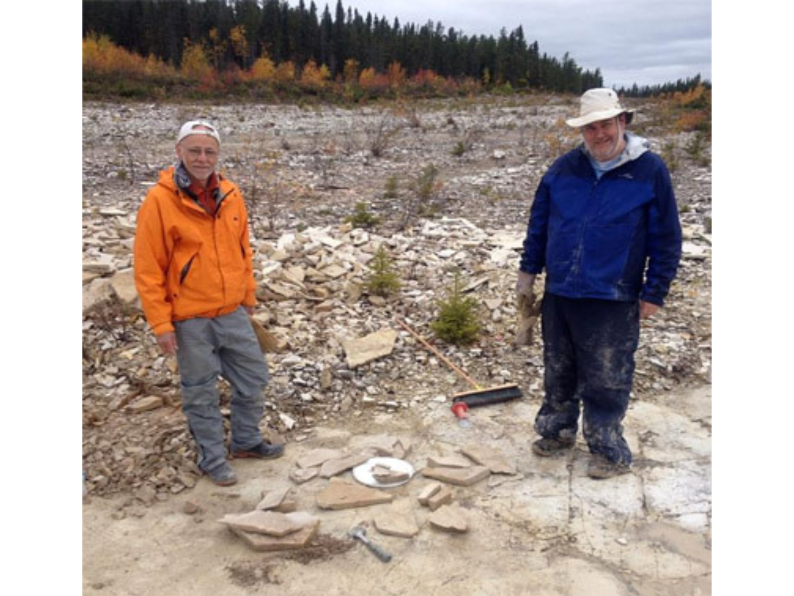 Two individuals wearing and orange and a blue jacket standing for a photo next to a small pile of rock slabs along with tools like a broom and hammer.