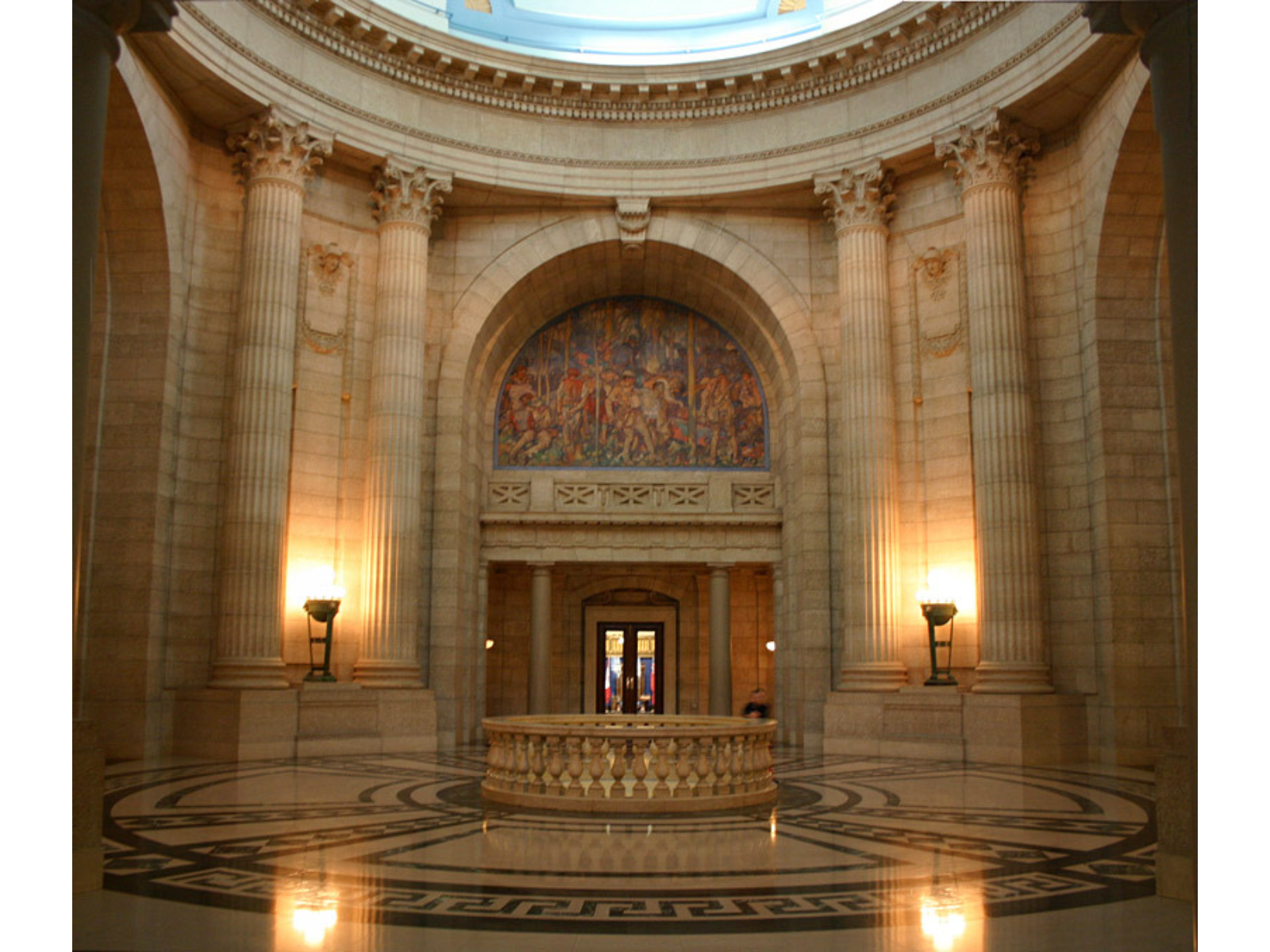 Interior of an imposing round room with pillars and a large arched doorway. In the centre of the room is a circular banister with a skylight looking down to the lower floor.