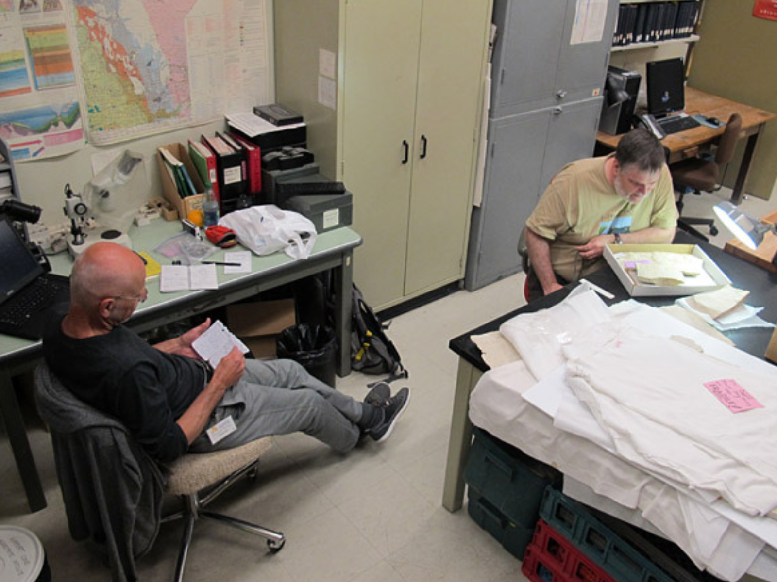 Two individuals each working at desks. One looks through paperwork while the other examines a specimen in a collections box.