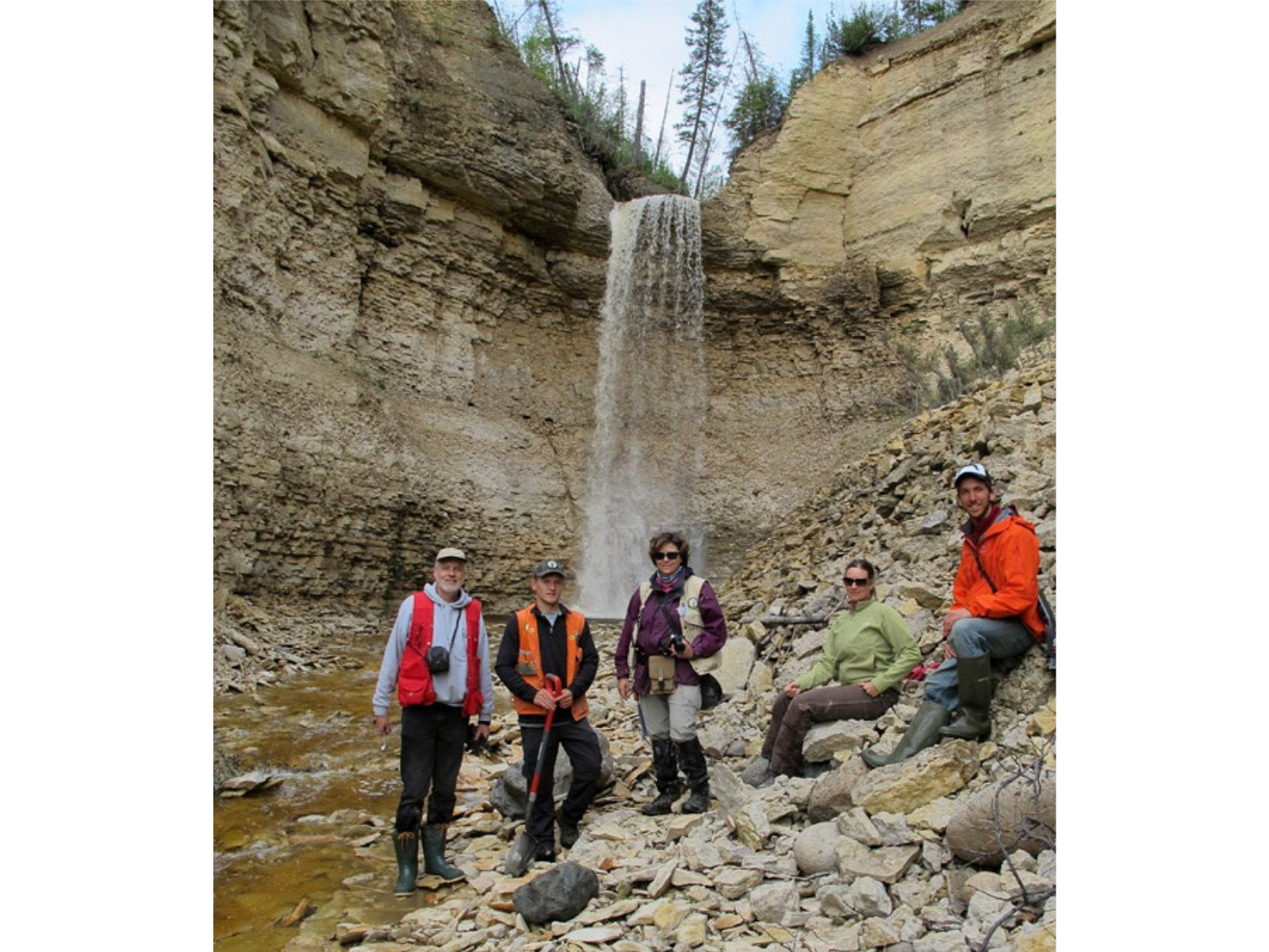 Five people posing together in front of a waterfall on the rocky shore of a creek.