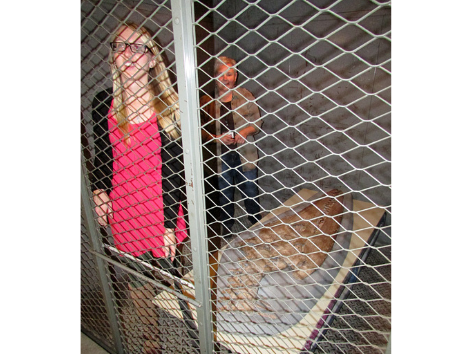 Two smiling individuals standing beside a cart containing a mounted plesiosaur skull in the metal cage of a freight elevator.