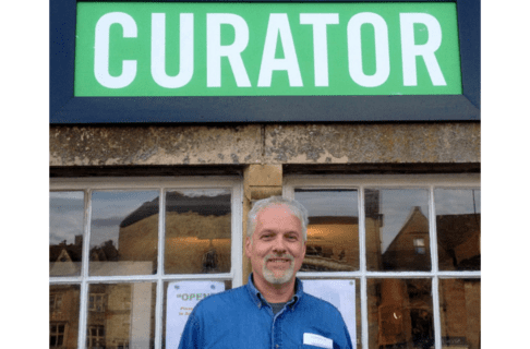 A smiling man, Graham Young, standing outside in front of a store under a green sign reading, "CURATOR".