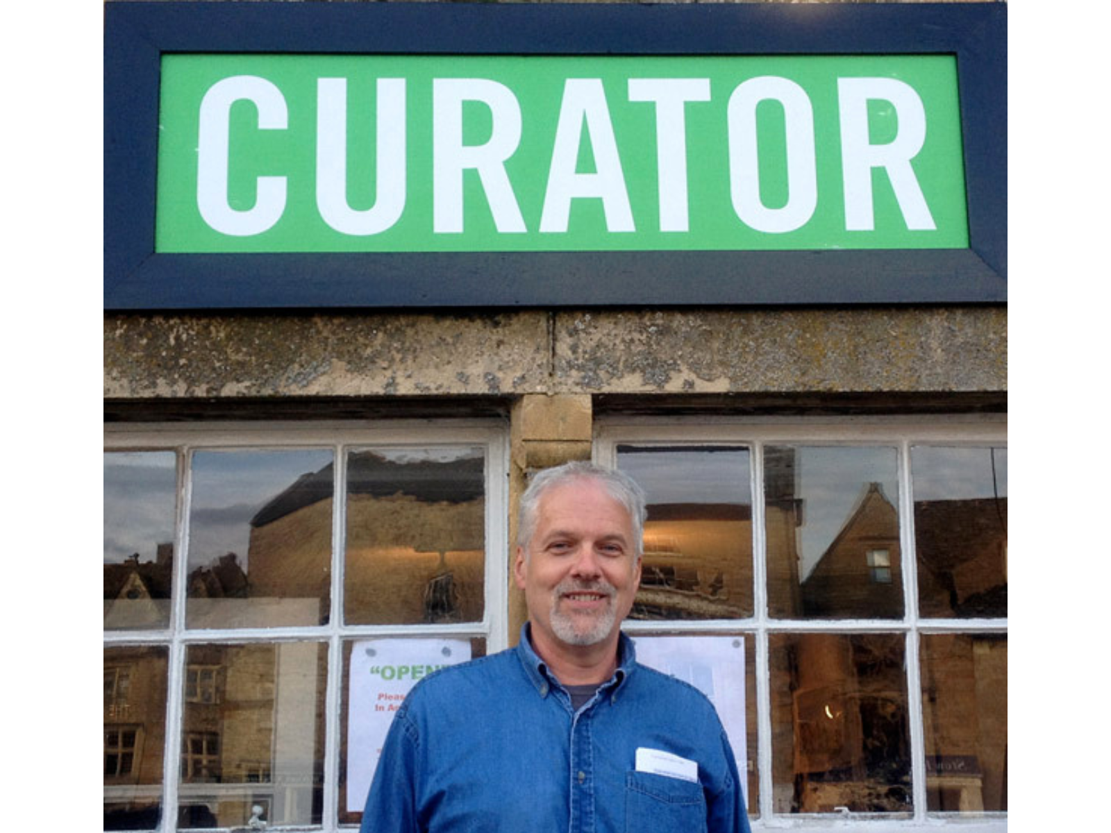 A smiling man, Graham Young, standing outside in front of a store under a green sign reading, "CURATOR".