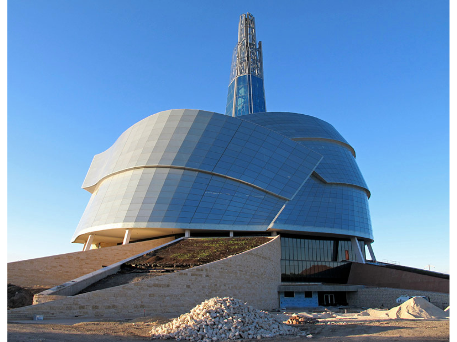 A large irregularly shaped building that is almost shaped like an upward growing spiral building to a central tower. This side of the building is primarily windows. In front of the building are piles of gravel and stone, as the site is under construction.