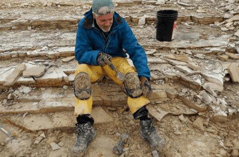 Dr. Graham Young seated on a rocky surface using a trowel to scrape thick mud off of knee pads worn over his yellow overpants. Mud covered his boots.