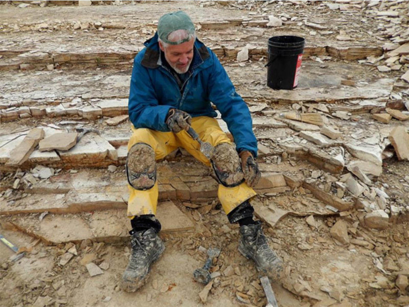 Dr. Graham Young seated on a rocky surface using a trowel to scrape thick mud off of knee pads worn over his yellow overpants. Mud covered his boots.