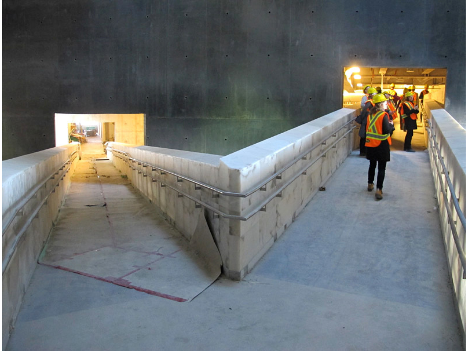 Standing in a corner of a ramp that goes lower to the left and rises to the right. Individuals in high-vis vests and hard hats are further up on the right-side ramp.