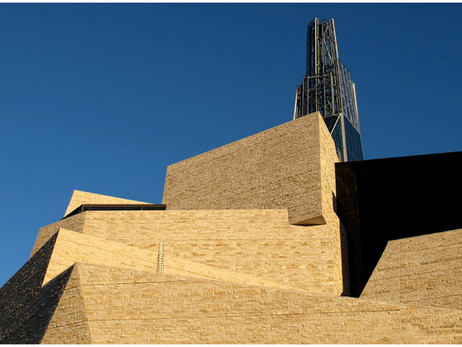 Looking up at the angular side of a stone building topped with a metal and windowed tower.