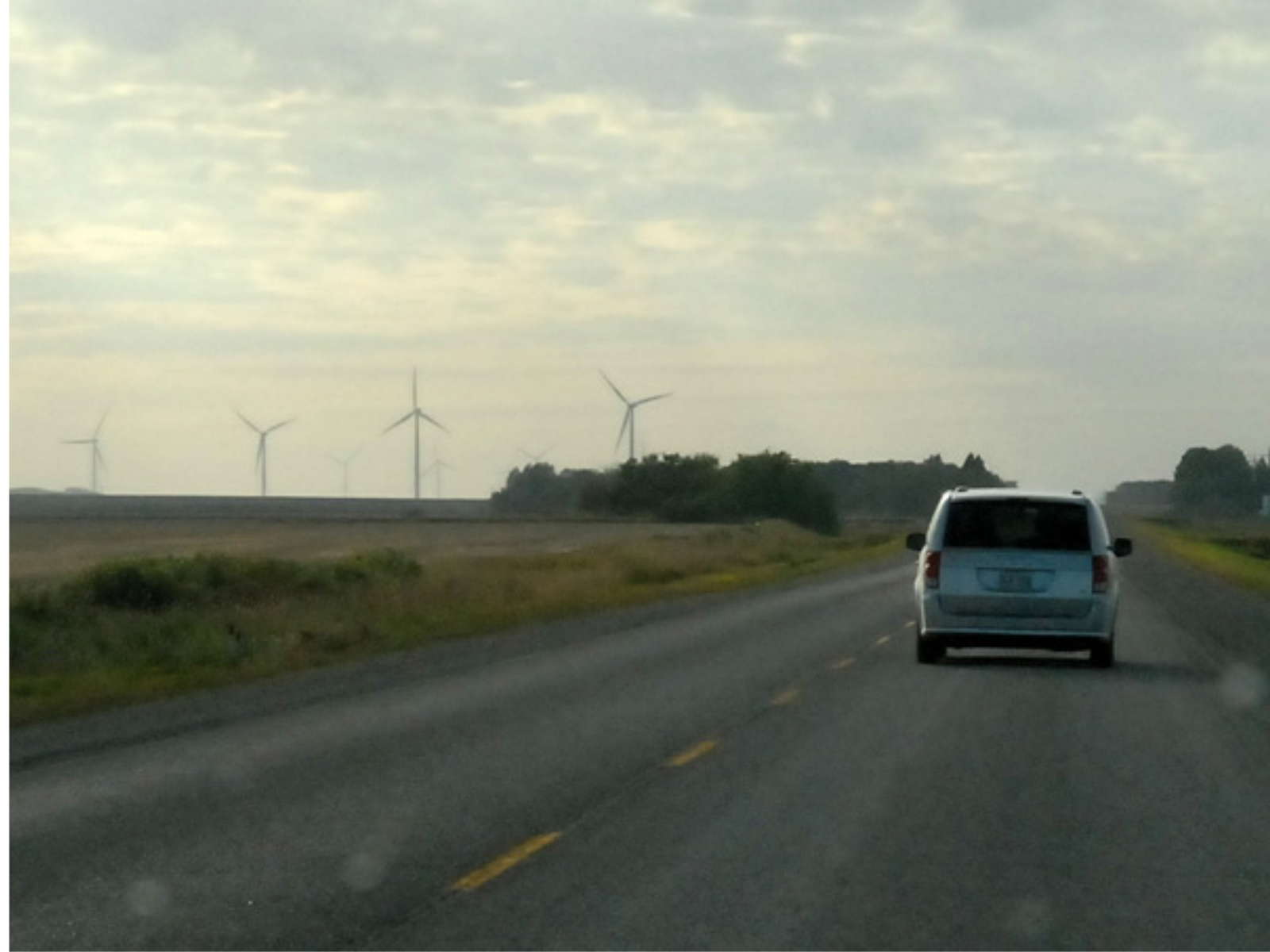 Photo taken from a moving vehicle on a single lane road. A vehicle drives in front, and in the distance, further in on the field, are three modern windmills.