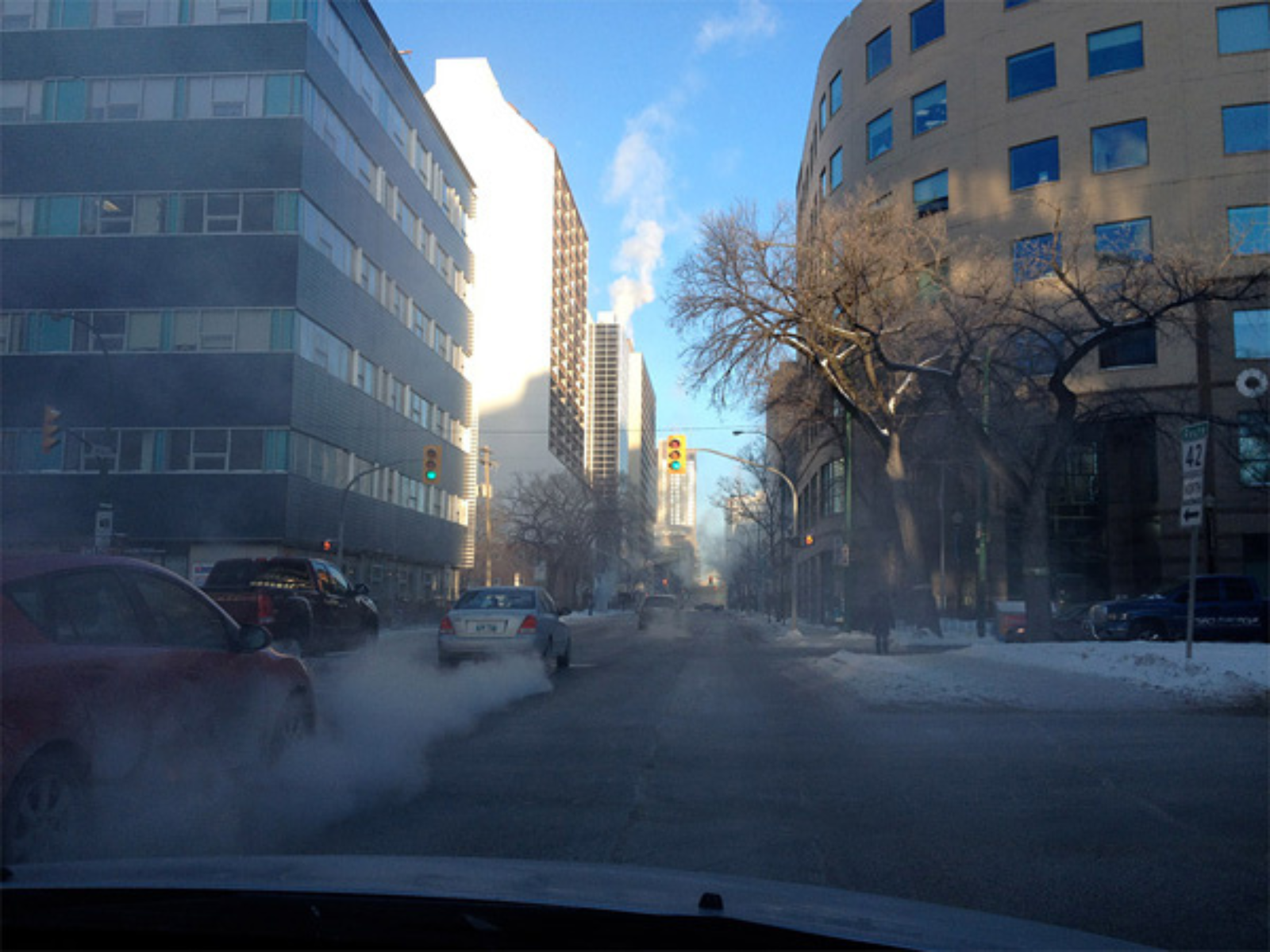 View down a downtown road between tall office buildings during winter. Under a blue sky there is snow along the sides of the road and leafless trees.