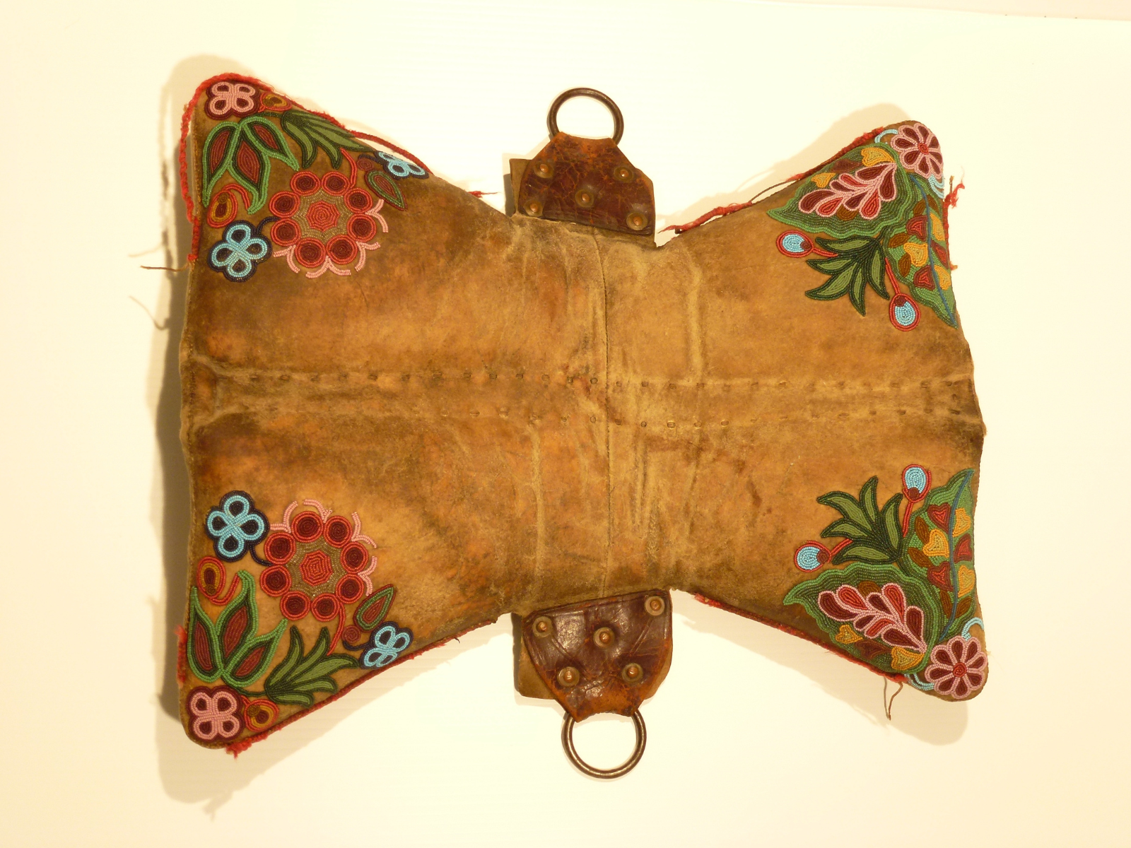 A leather padded saddle with beaded floral detailing on the four corners and clasps at either end of the centre.