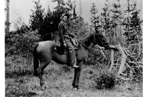 A black and white photo of an individual on horseback in front of a wooded area.