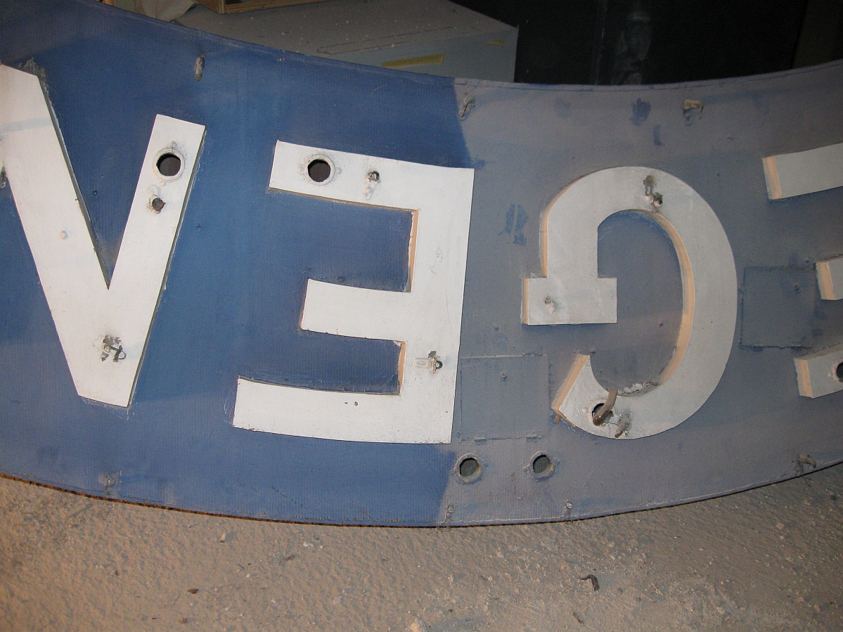 A portion of a large sign, half vacuumed. One half is very dimmed with dust, the other is clean and blue with white lettering.