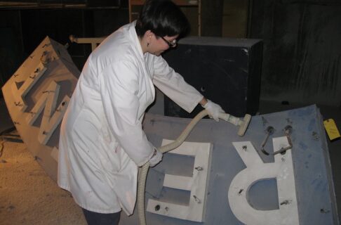 An individual wearing a white lab coat holding the hose and nozzle of a vacuum cleaner to a large dusty sign.