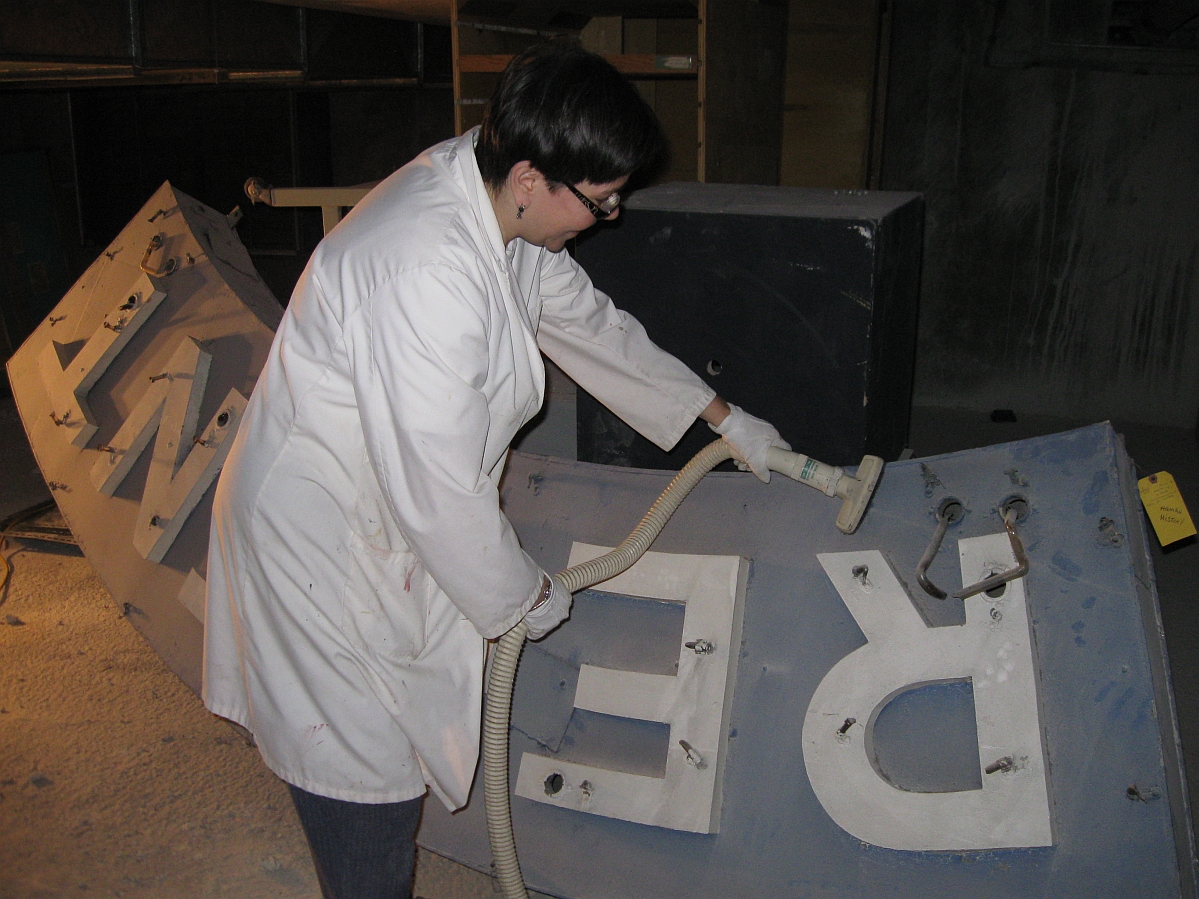 An individual wearing a white lab coat holding the hose and nozzle of a vacuum cleaner to a large dusty sign.