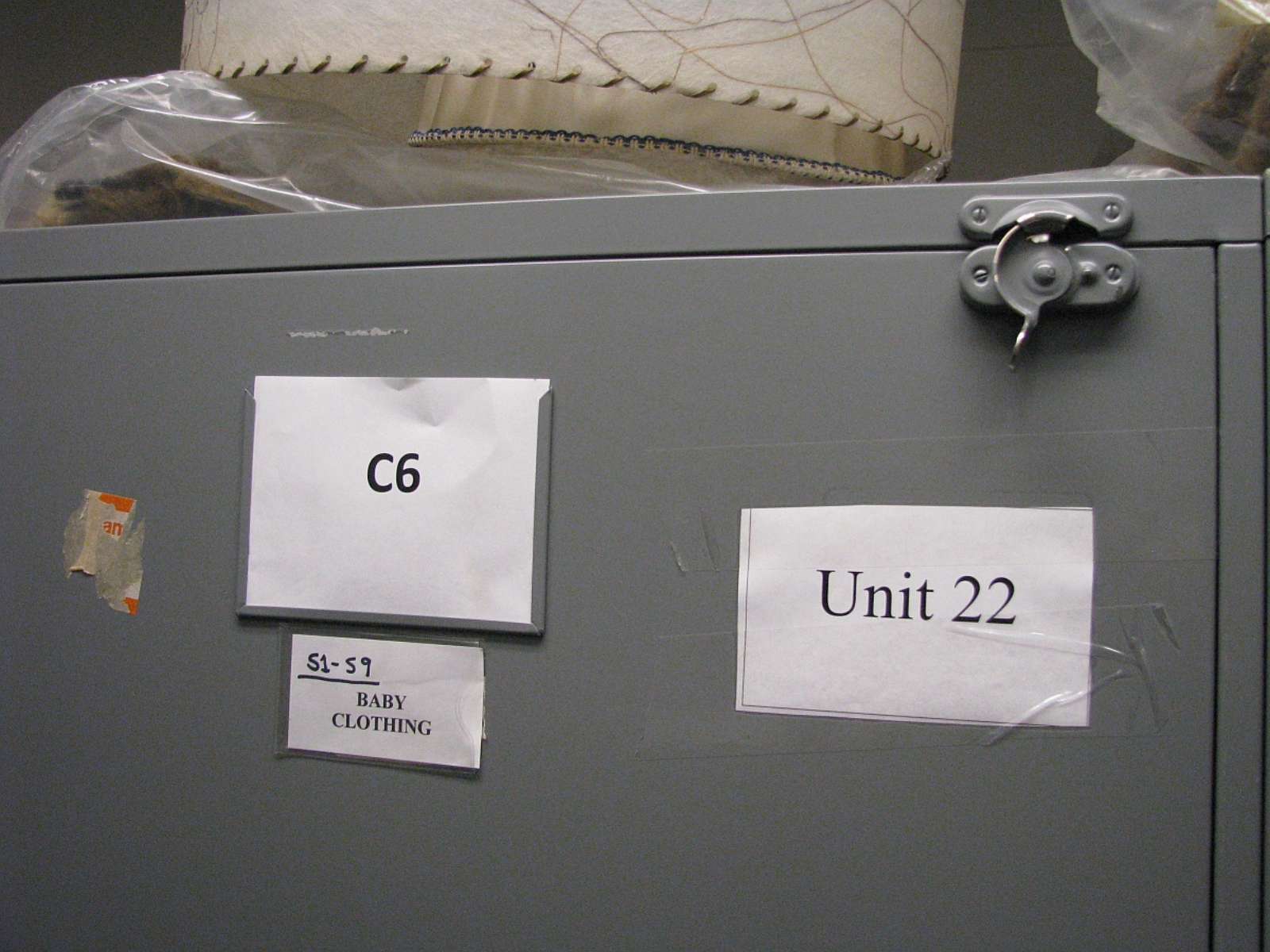The door of a closed storage cabinet with a number of white labels reading, "Unit 22", "C6", and "51-59 / Baby Clothing".