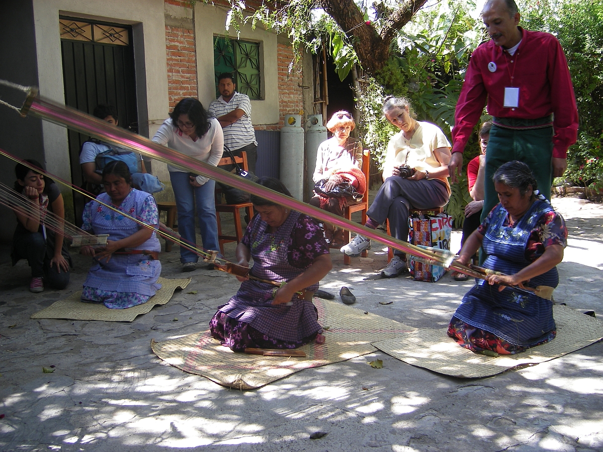 Three women kneeling on mats hold the ends of long weaving threads and looms. Behind them a number of people are seated or standing watching.