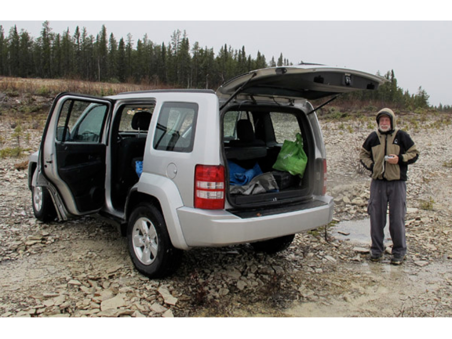 A man wearing a coat and hood, with one hand in his pocket and the other holding a camera, stands on a rocky wet surface next to the open trunk of an SUV.