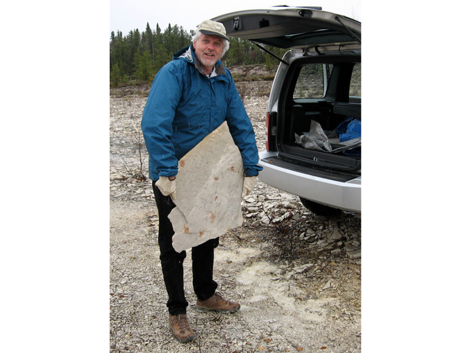 Dr Graham Young, wearing a blue jacket and a baseball cap stands at the open trunk of a car holding a large stone slab with a fossil embedded in it.