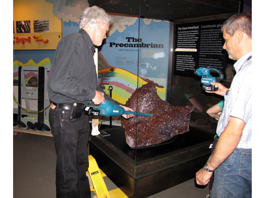 Two individuals stand next to a large amethyst in a case. One is holding a hand-held vacuum, cleaning the amethyst, as the other holds up a work light.
