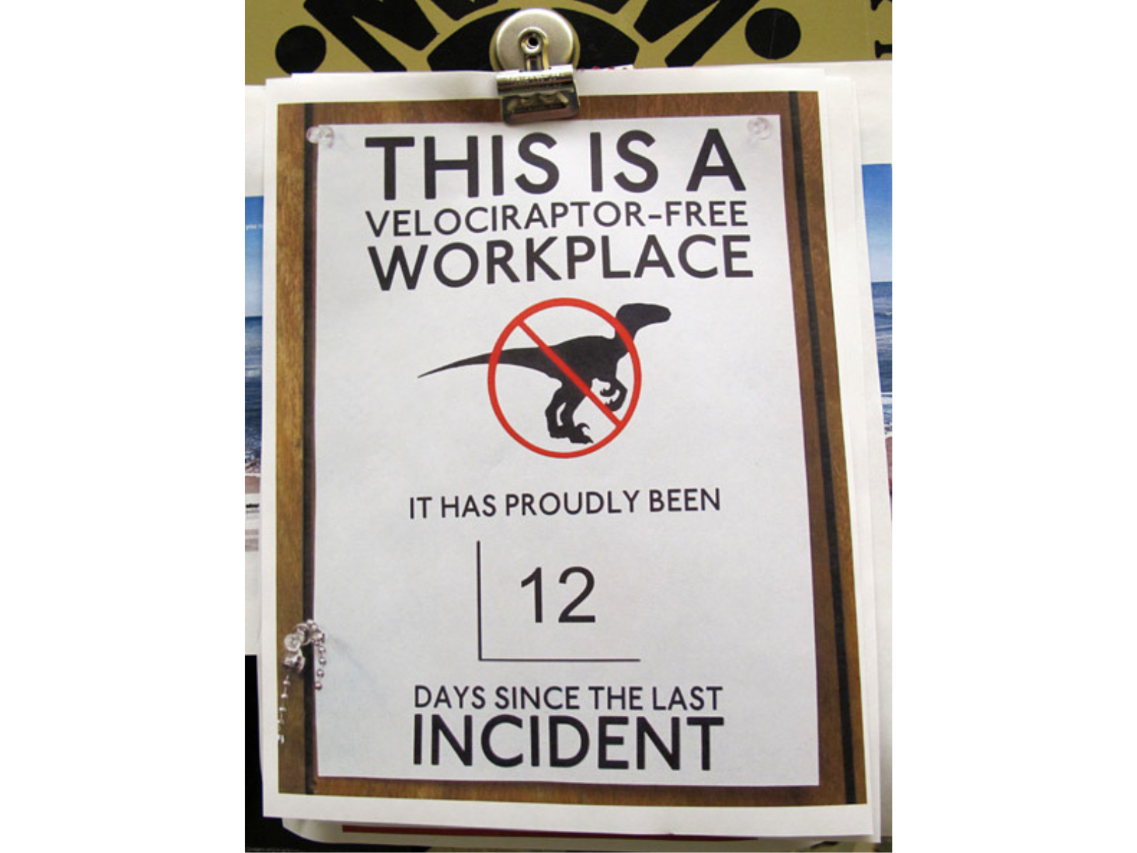 A printed sign showing a silhouette of a velociraptor with a circle around it and a line striking through. Text reads, "This is a velociraptor-free workplace / it has proudly been / 12 / days since the last incident".