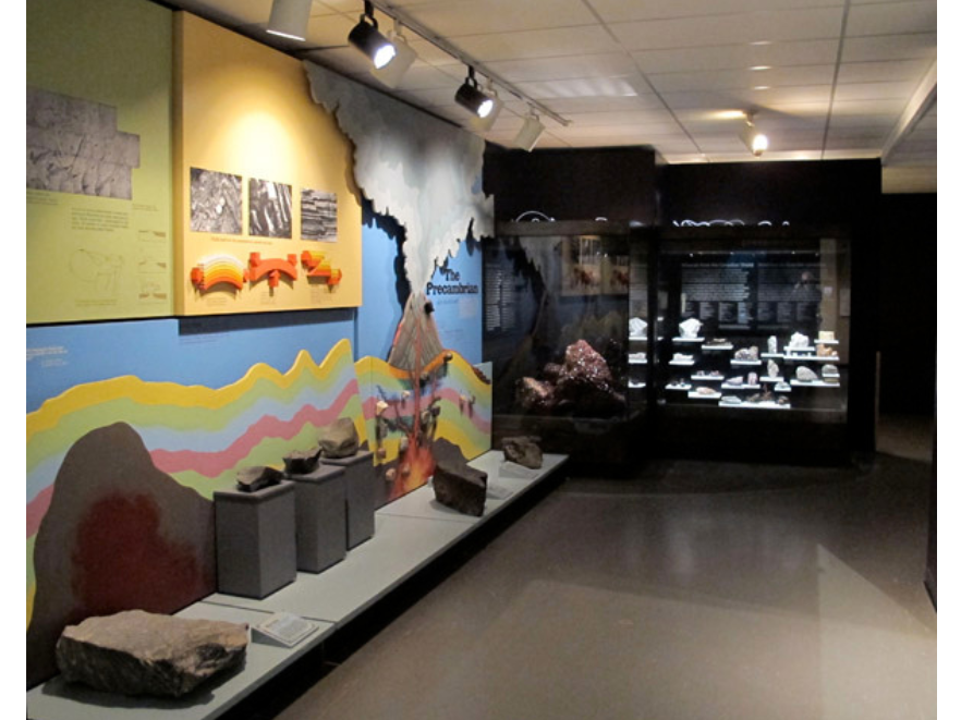 A view of the entrance way into the Earth History Gallery, with a exhibit of the layers of the earth along the left side wall, and a dramatically lit new display case at the far end showing a selection of mineral specimens.