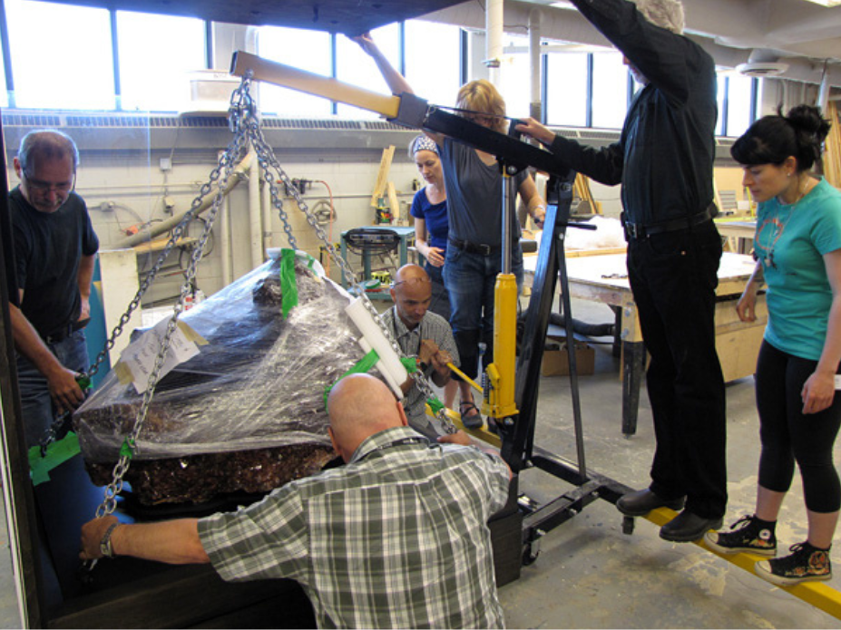 Seven people grouped around a hoist lifting a steel platform with a large wrapped rock on it. A case base has been moved under the lifted piece.