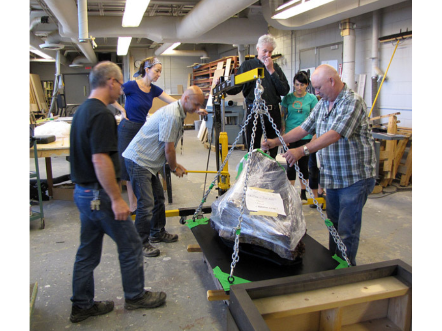 Six individuals group around a loading hoist that is lifting a steel platform with a large wrapped rock on it.