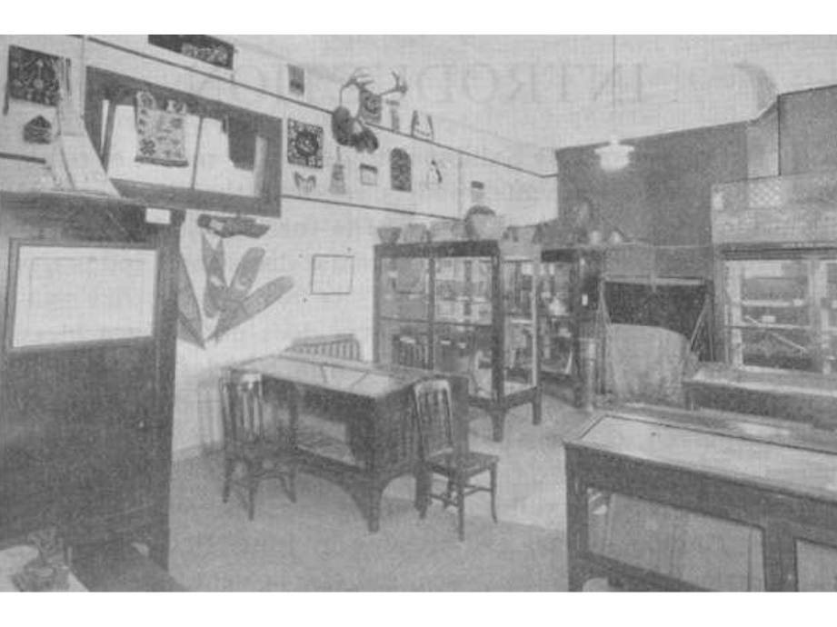 A black and white photograph of a room set up as a museum exhibit with display cases, display tables, and artifacts mounted on the walls.