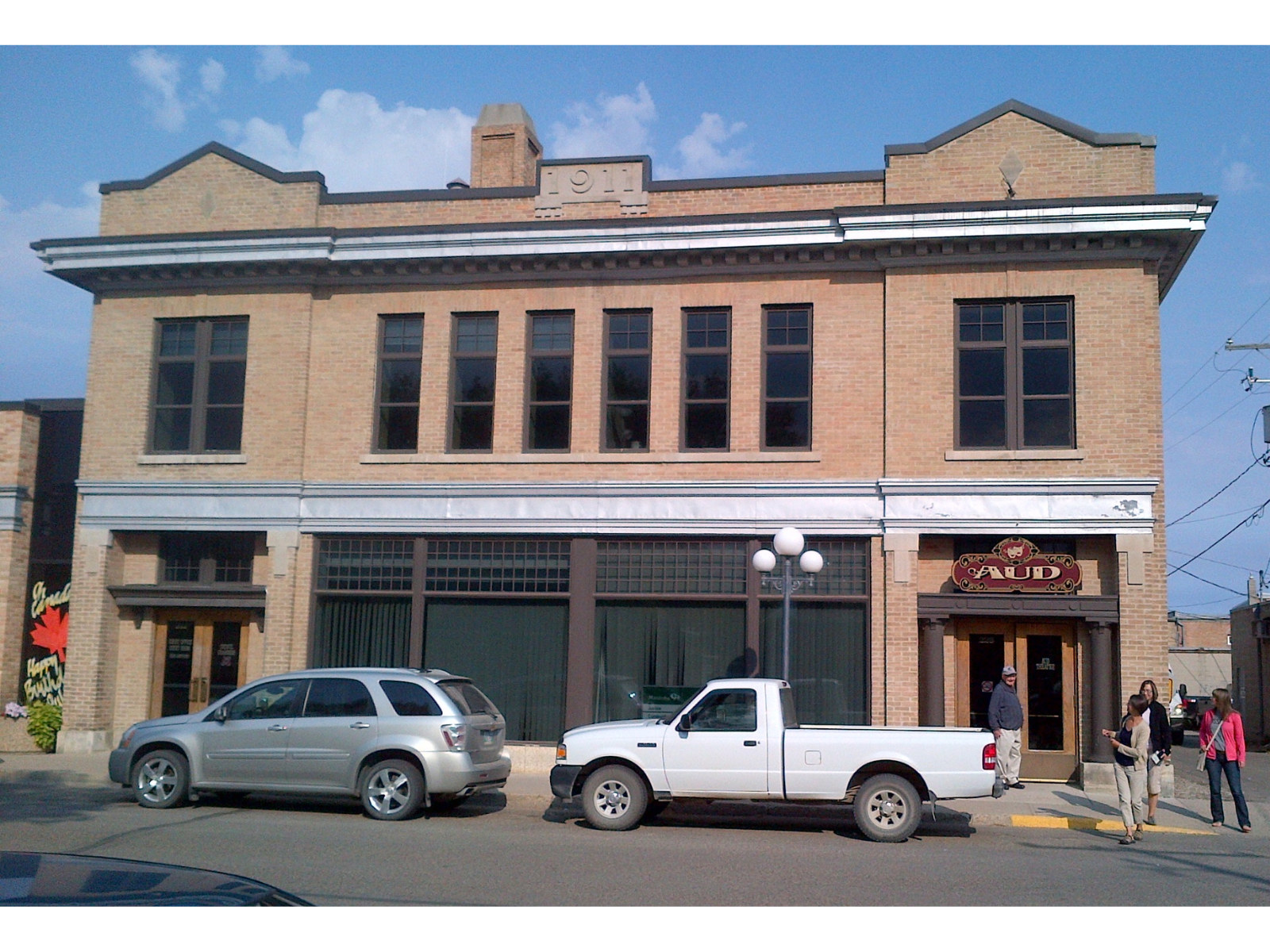 A two-storey reddish-brown brick building along a road. Above the entrance to the right is a sign reading "The Aud". Four individuals stand near each other by the door.
