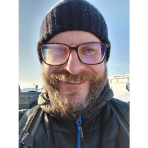 Selfie photo of a bearded man wearing a knit toque, thick rimmed glasses, and a fully zipped up dark parka standing outside under a blue sky.