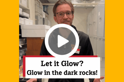 A screenshot of a video, an individual stands inside museum collection storage. There's a play button over the screenshot and overlaid text reads, "Let it Glow? Glow in the dark rocks!".