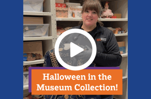 A screenshot of a video, an individual stands in the museum collections storage holding up a Halloween costume dress. There's a play button over the screenshot and overlaid text reads, "Halloween in the Museum Collection".