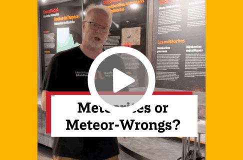 A screenshot of a video, an individual stands in the Science Gallery next to a meteor exhibit. There's a play button over the screenshot and overlaid text reads, "Meteorites or Meteor-Wrongs?".