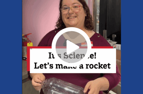 A screenshot of a video, an individual stands in the Science Gallery holding an empty 2 litre pop bottle. There's a play button over the screenshot and overlaid text reads, "It's Science! Let's make a rocket".