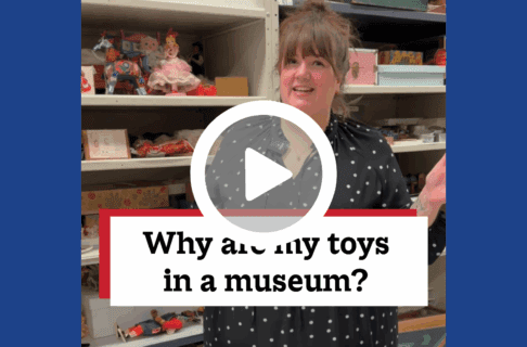 A screenshot of a video, an individual stands inside museum collection storage, next to shelves of toys. There's a play button over the screenshot and overlaid text reads, "Why are my toys in a museum?".